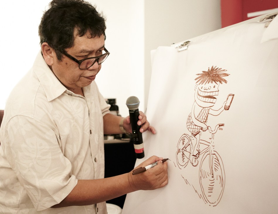 Cartoonist Lat draws a character on a clipboard while holding a microphone at a festival event.