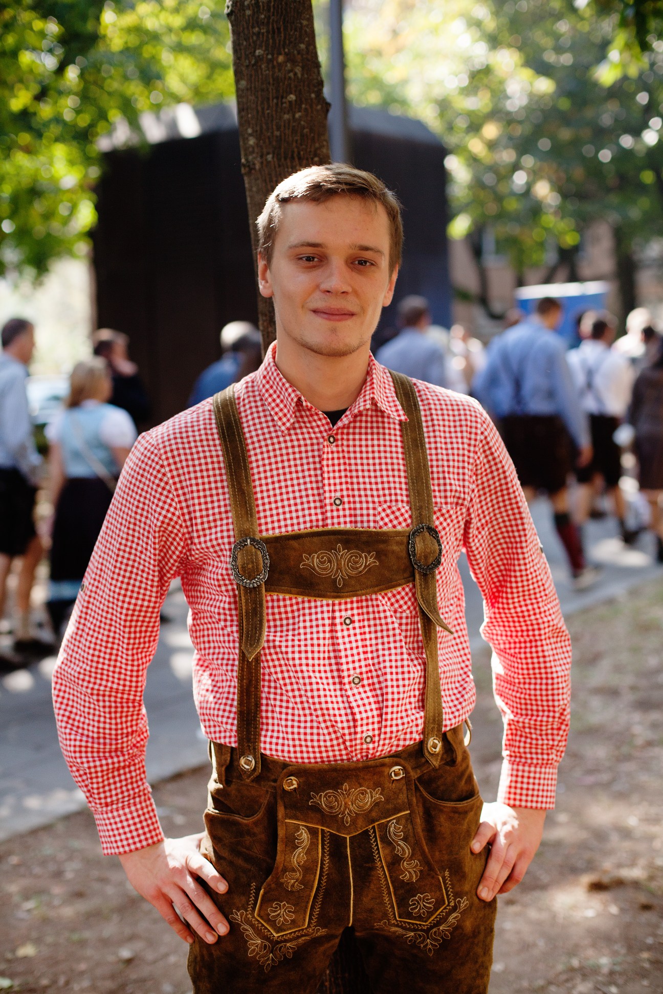 Young man wearing traditional Bavarian lederhosen with embroidered brown suede shorts, braces, and red gingham shirt at Oktoberfest.