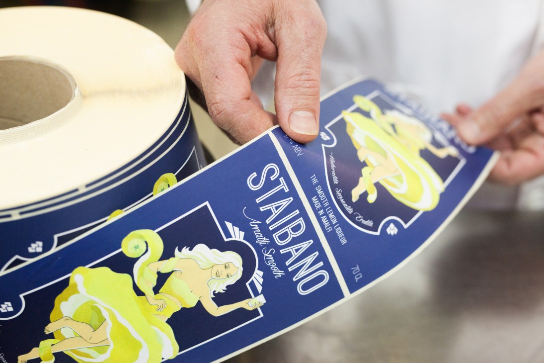 Hand applying a blue and yellow Staibano lemon liqueur label to a bottle during manual labeling process.