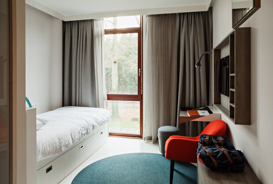 Modern student bedroom with bed, desk, orange chair, and forest view through glass door.