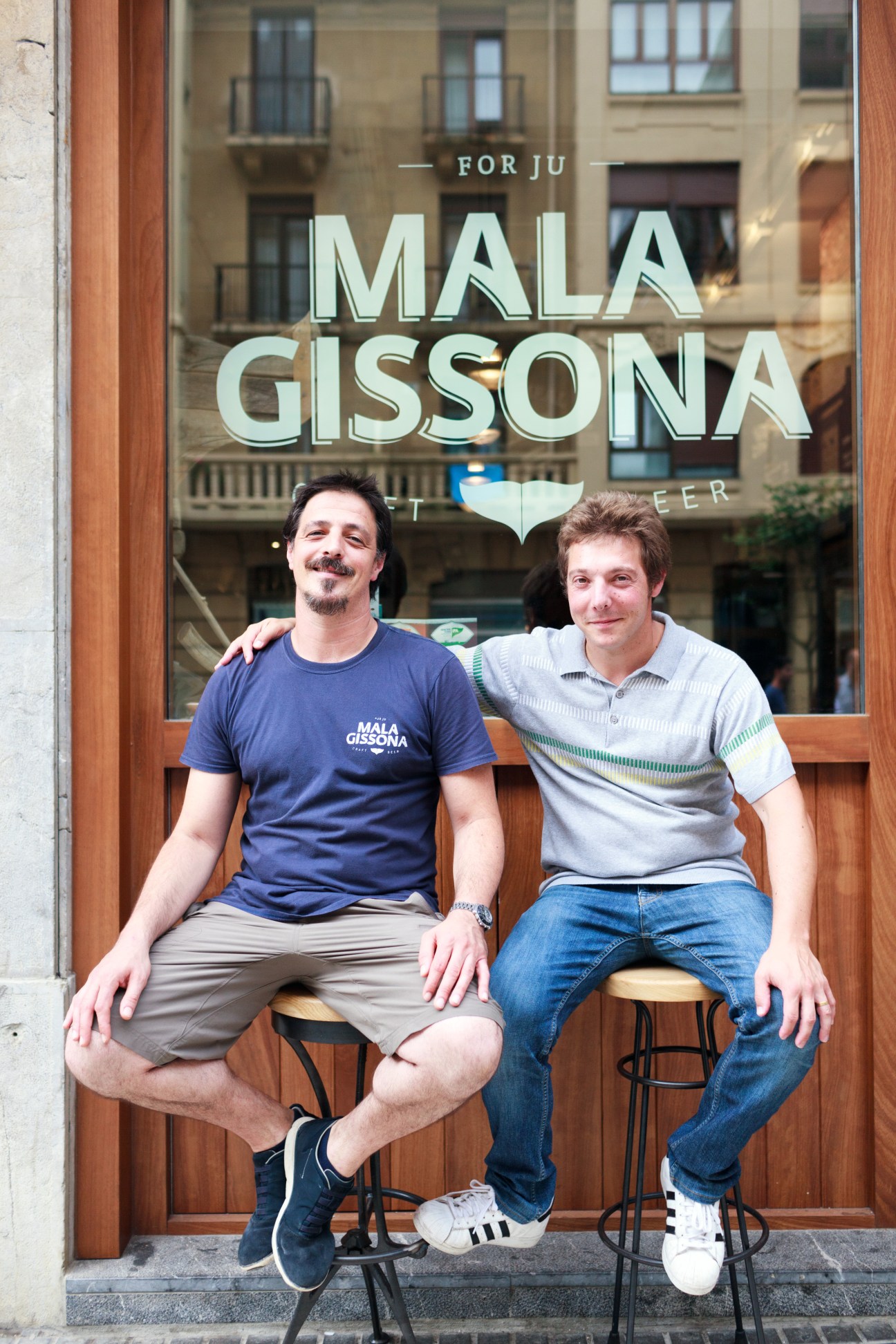 Two men sit on stools outside Mala Gissona, a beer house in San Sebastián, Spain, with the storefront sign visible behind them.