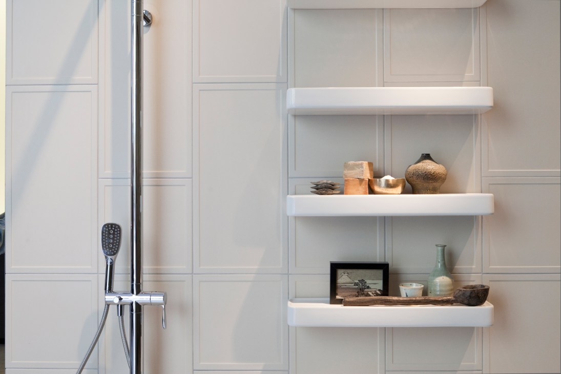 Modern bathroom with white paneled walls, chrome shower fixture, and white floating shelves displaying decorative objects.