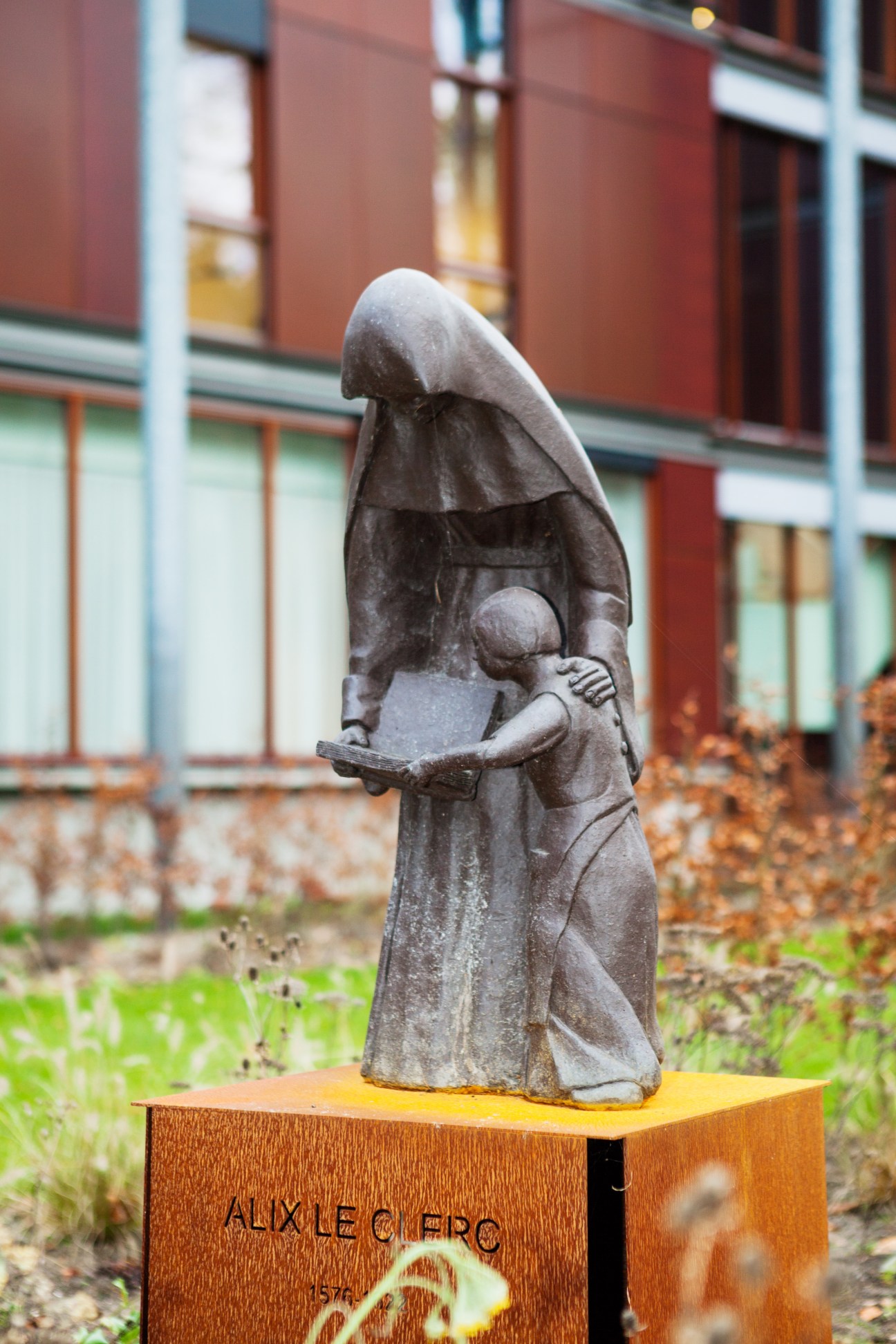 Stone statue of a nun in habit holding a book, mounted on a wooden plaque labeled 'AUXLECRO' in front of a red brick building.