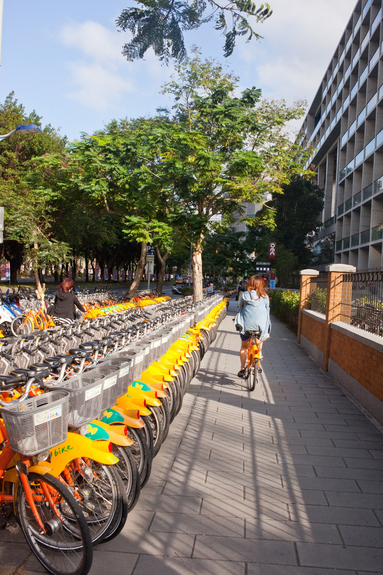 Woman riding yellow and orange bike past rows of parked U-bikes at National Taiwan University station.