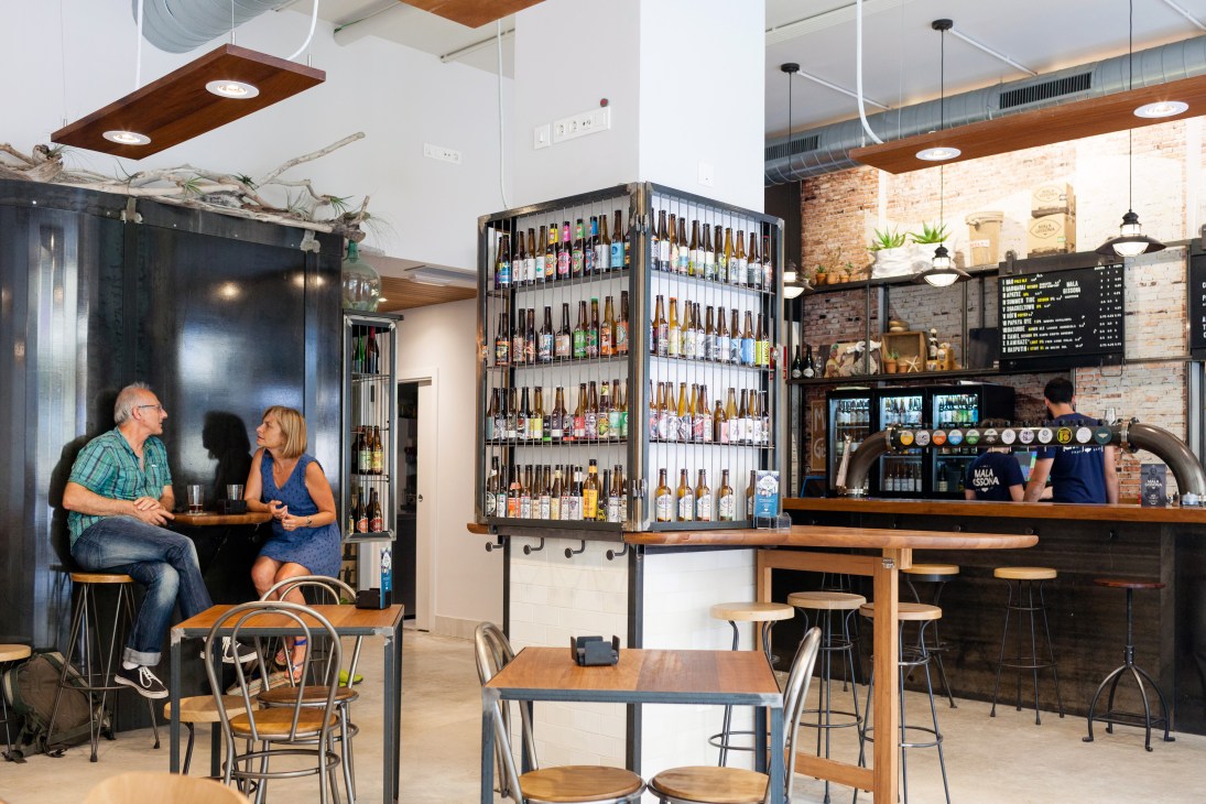 Interior of Mala Gissona beer house with customers seated at tables and a large illuminated beer bottle display behind the bar.