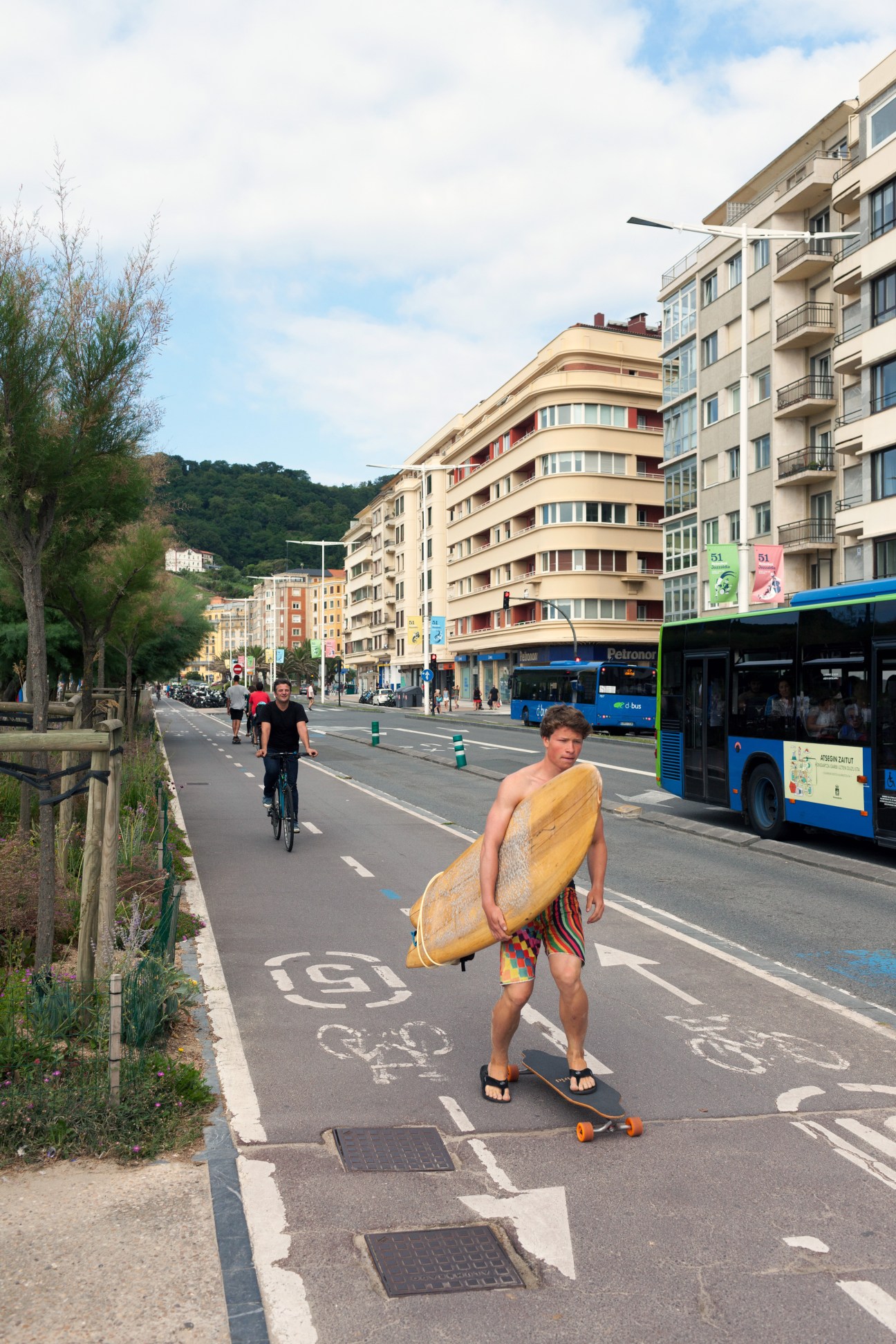 A surfer carries a yellow surfboard down a pedestrianized street in San Sebastián's Gros neighbourhood, passing cyclists and a blue bus.