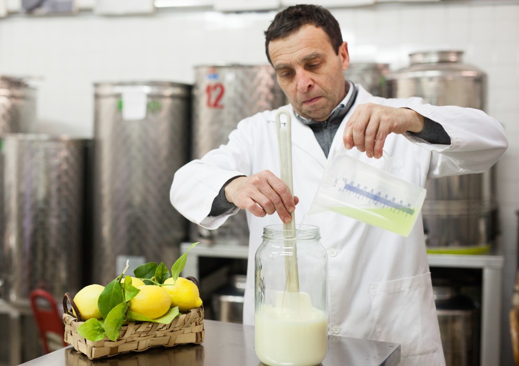 Man in white lab coat pouring lemon liqueur into a glass jar in a factory laboratory with steel tanks in background.