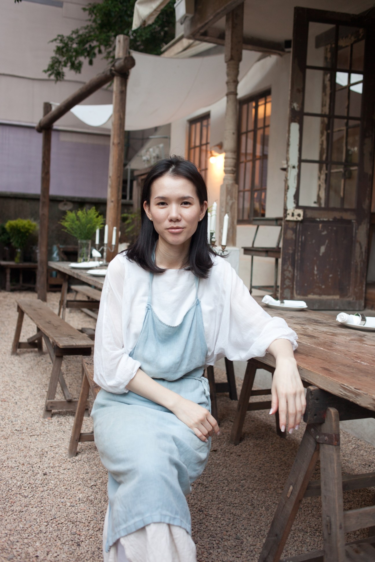 Zoe Yang, owner of restaurant Xiang Se, sits in a courtyard wearing a light blue slip dress over a white shirt.