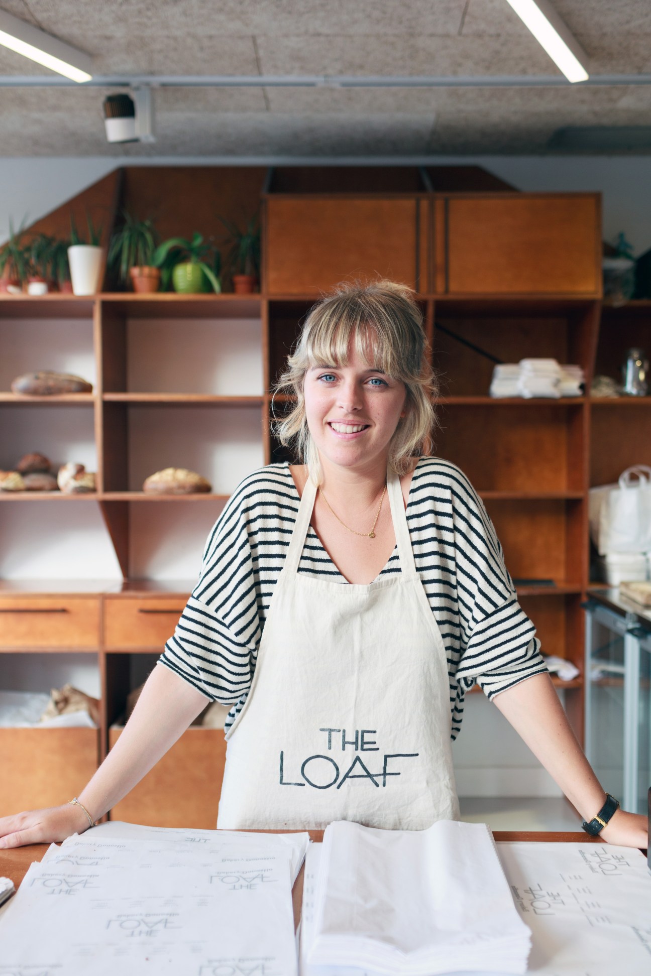 Woman wearing striped shirt and The Loaf apron smiling at counter in bakery with wooden shelving behind her.
