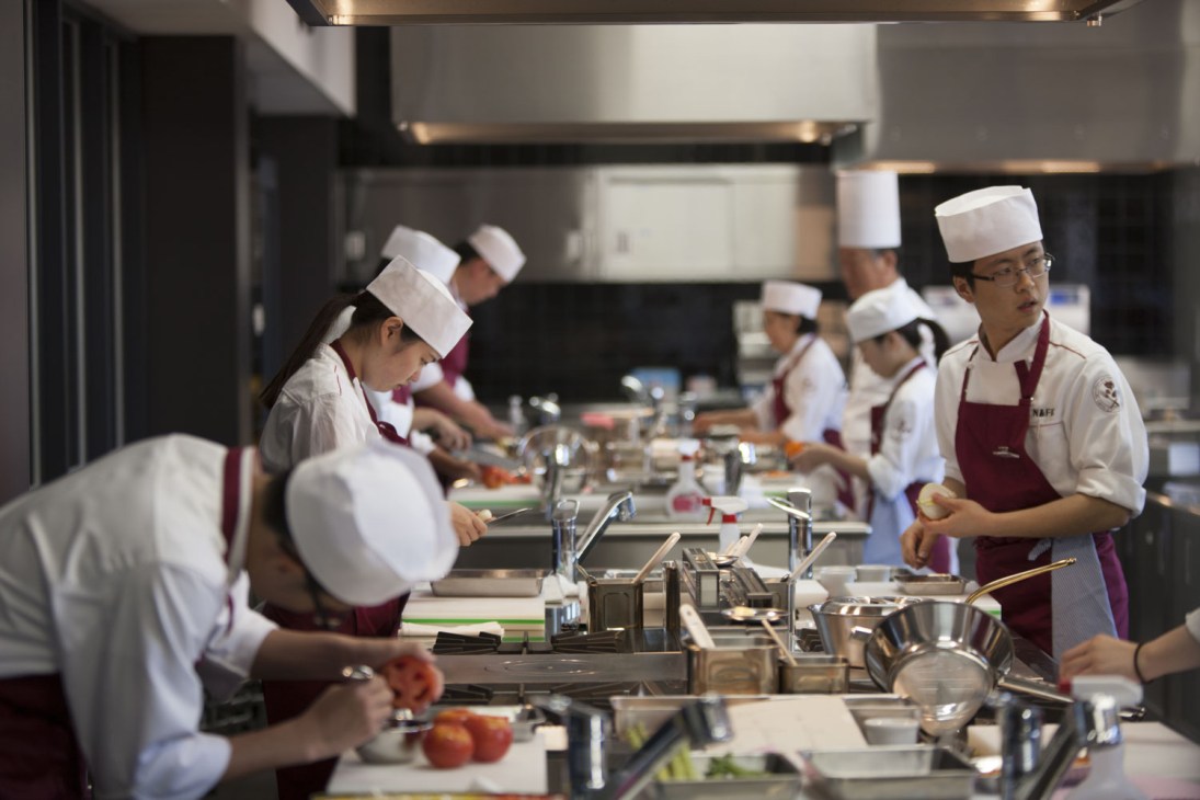 Students in white chef uniforms and hats work at cooking stations in a modern kitchen classroom.