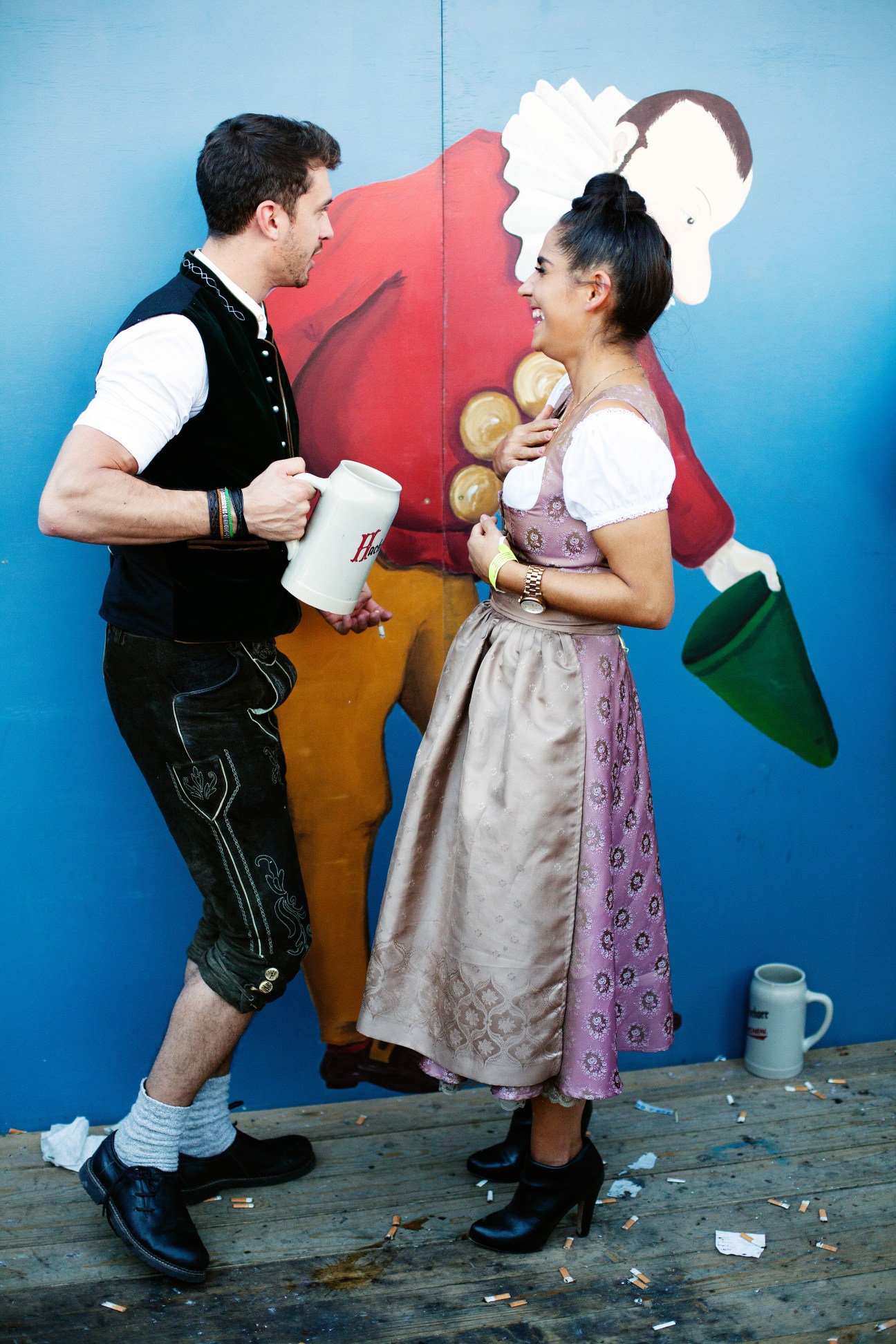 A couple in traditional Bavarian tracht clothing holding beer mugs and pretzels against a blue backdrop.