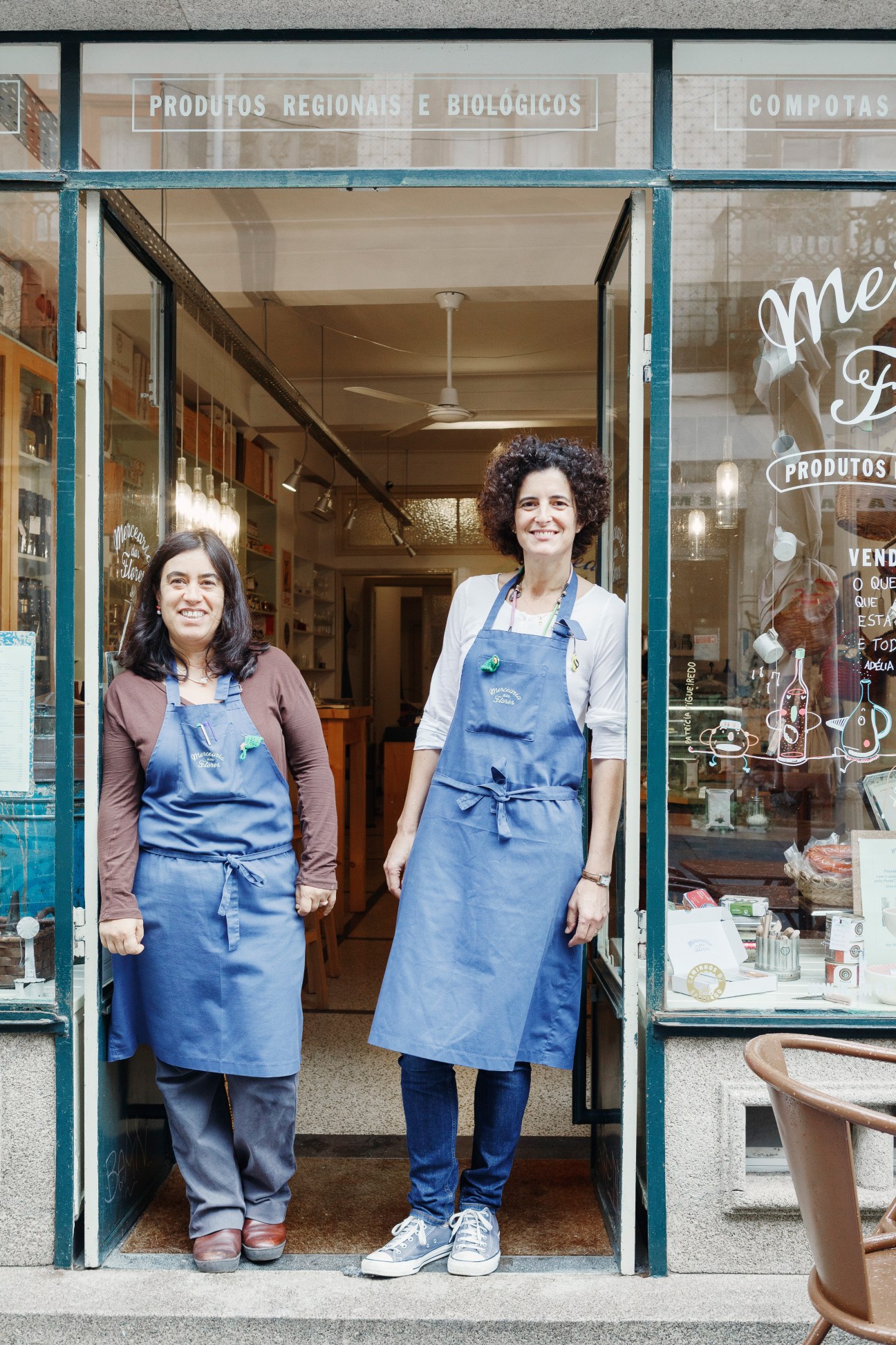 Two women in blue aprons stand in the doorway of Mercearia das Flores, a Portuguese gourmet grocery store in Porto.