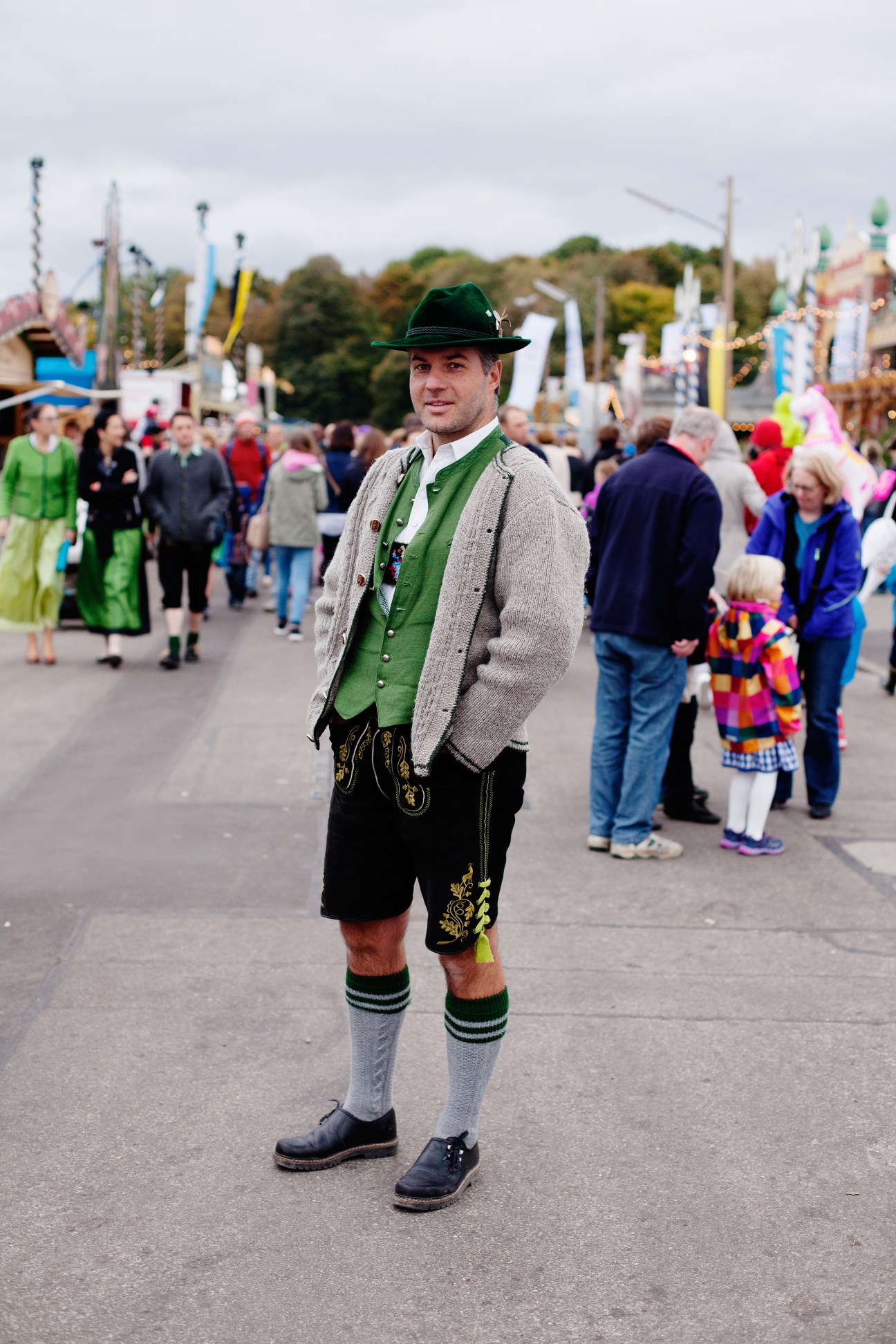 Man wearing traditional Bavarian tracht with green vest, black lederhosen, grey cardigan and green hat at Oktoberfest.