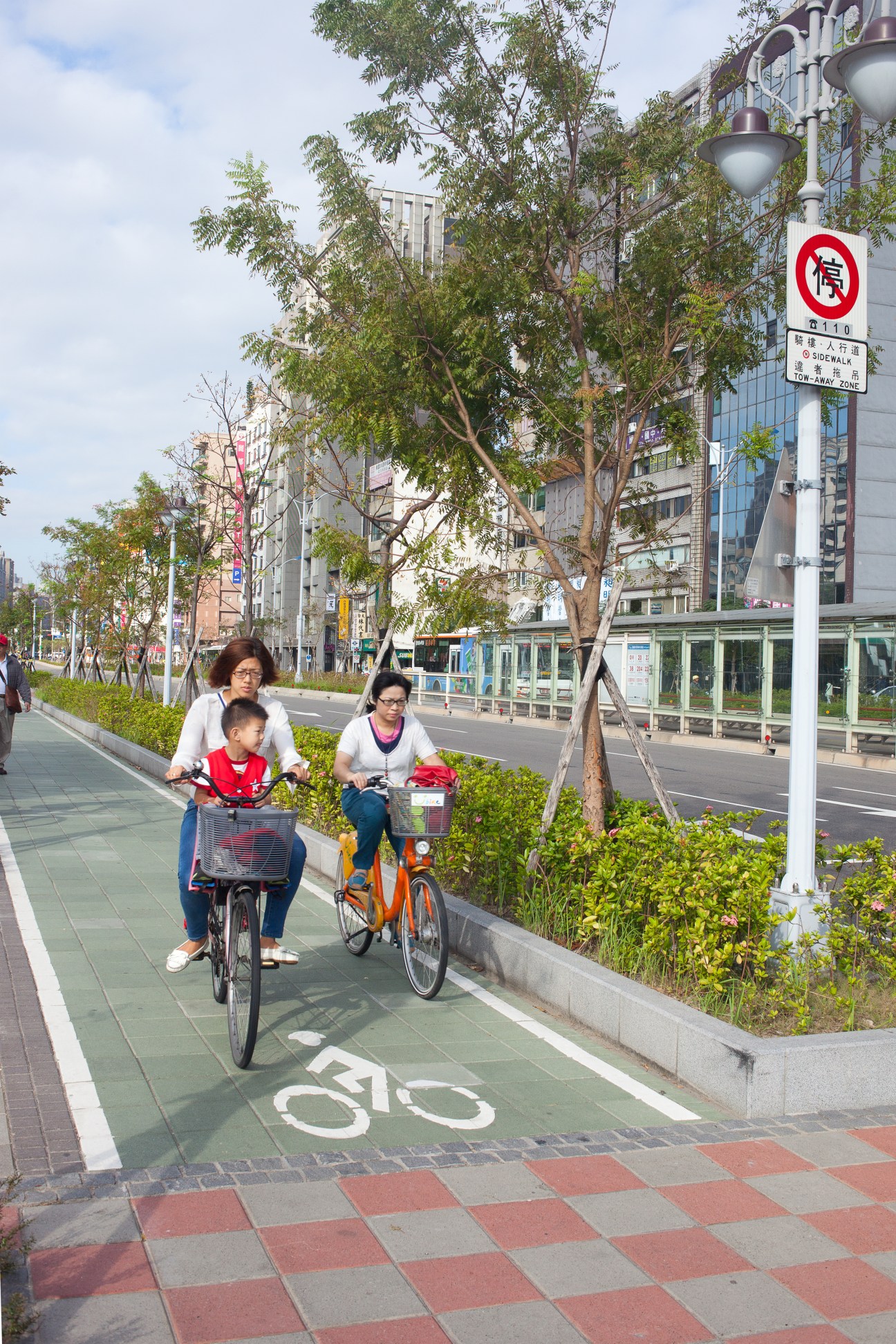 Two women and a child ride bikes on a dedicated cycle lane in Taipei with trees and urban buildings in the background.