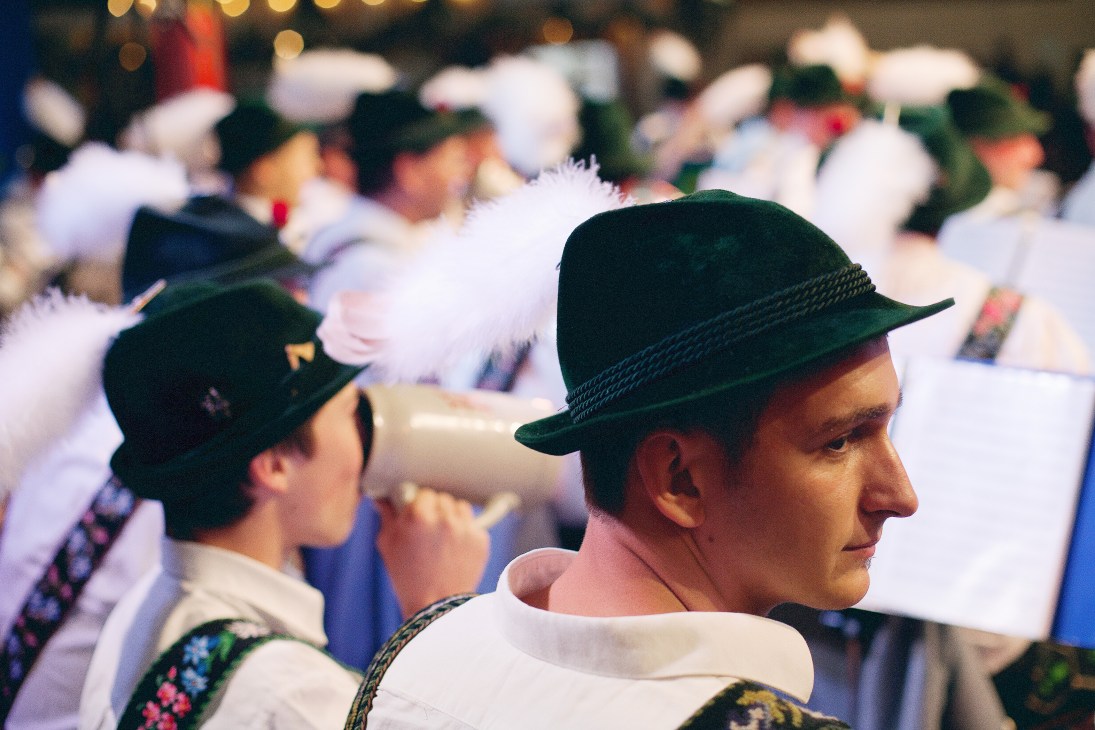 Men wearing traditional Bavarian tracht with dark green hats and embroidered vests sit in the Herzkasperl beer tent at Oktoberfest.