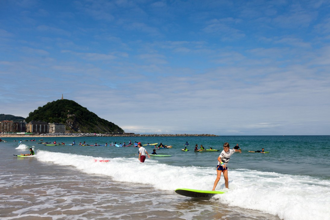 Surfers and swimmers in the ocean at Zurriola Beach in San Sebastián with a green forested hill visible in the distance.