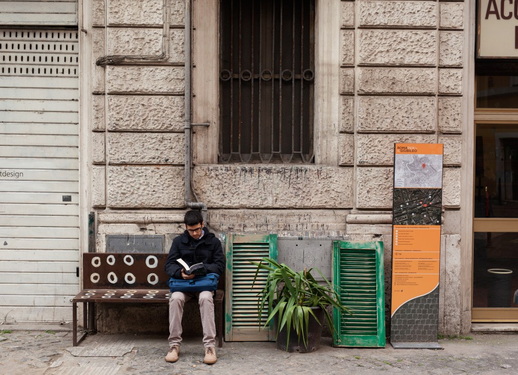 Man reading on a bench in Piazza della Suburra, Rome, against weathered ochre building facade.