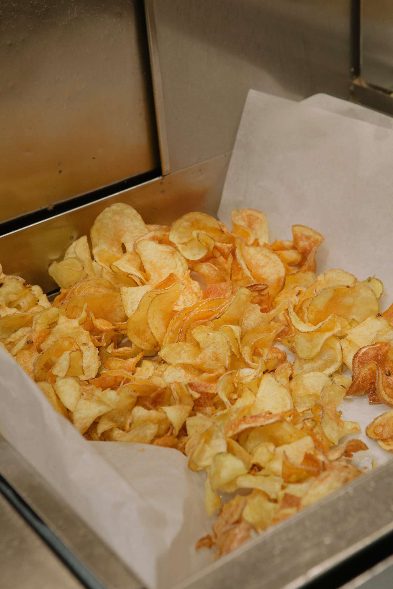 Golden potato crisps fresh from the fryer on white paper in a metal tray.