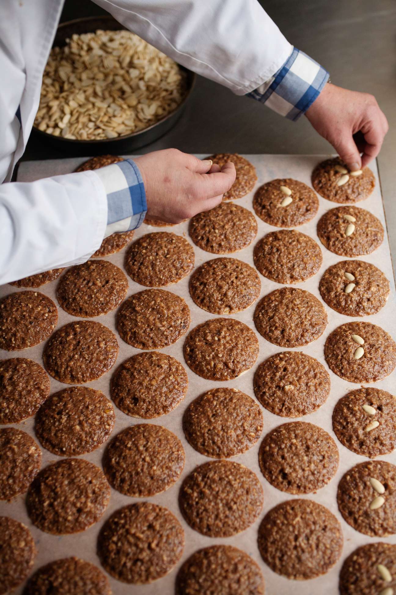 A baker in white chef's coat arranging gingerbread cookies with almond pieces on parchment paper.