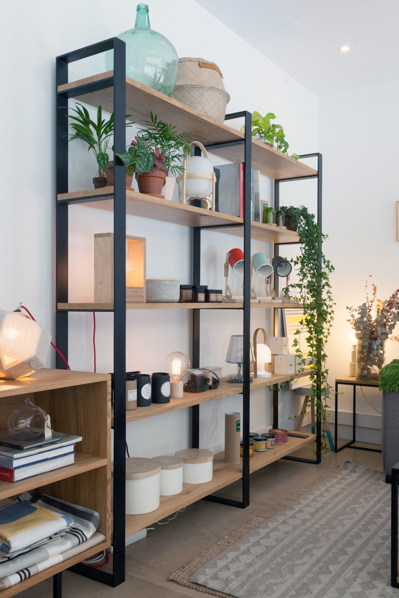 Industrial shelving unit with black metal frame and wooden shelves displaying plants, home décor items, and lighting fixtures.