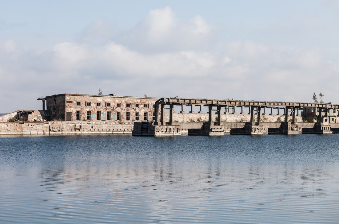 Abandoned Soviet-era waterfront facility with deteriorating concrete structures reflected in calm water under cloudy sky.