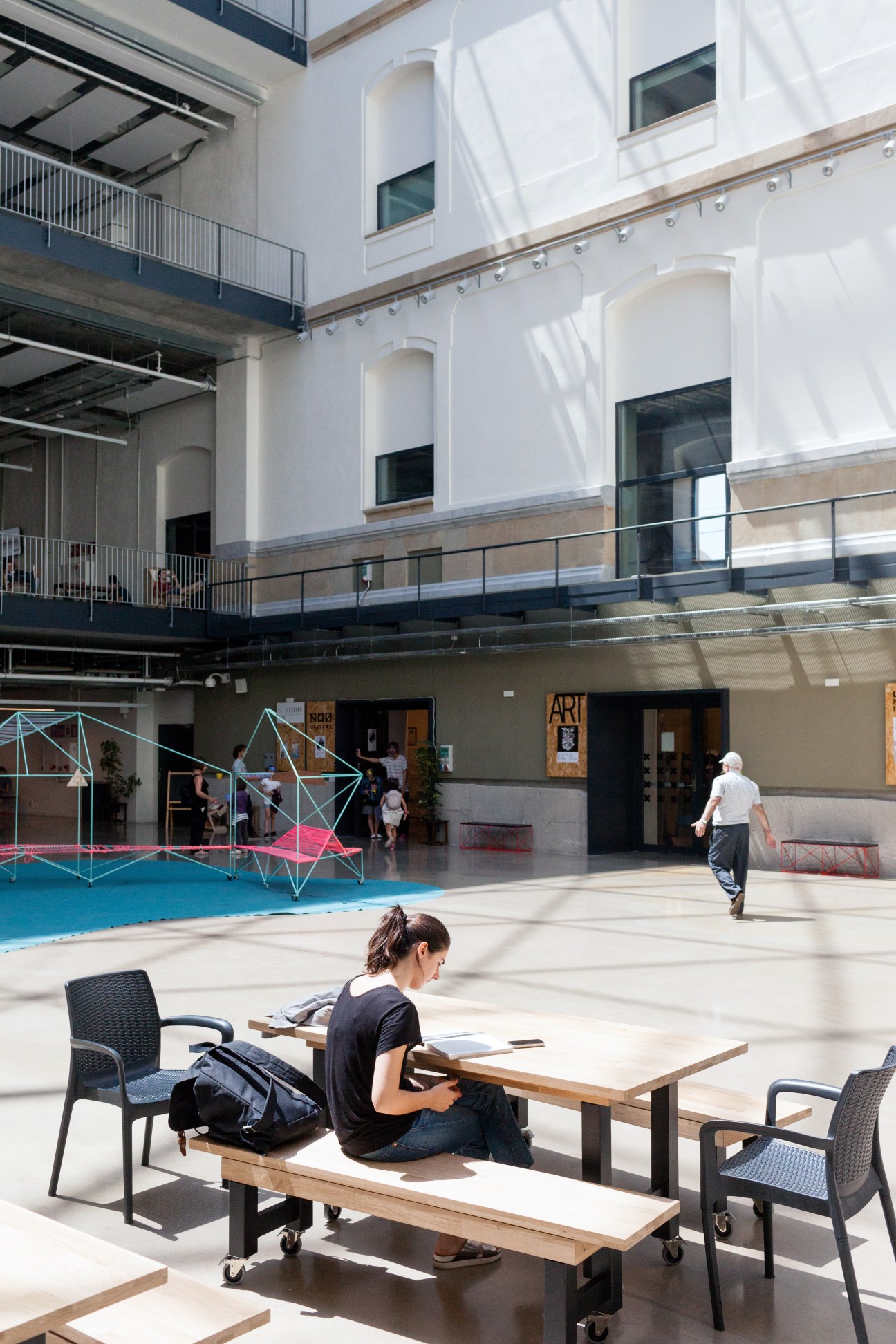 Woman sitting at wooden table in bright courtyard with white building facade and geometric art installation.