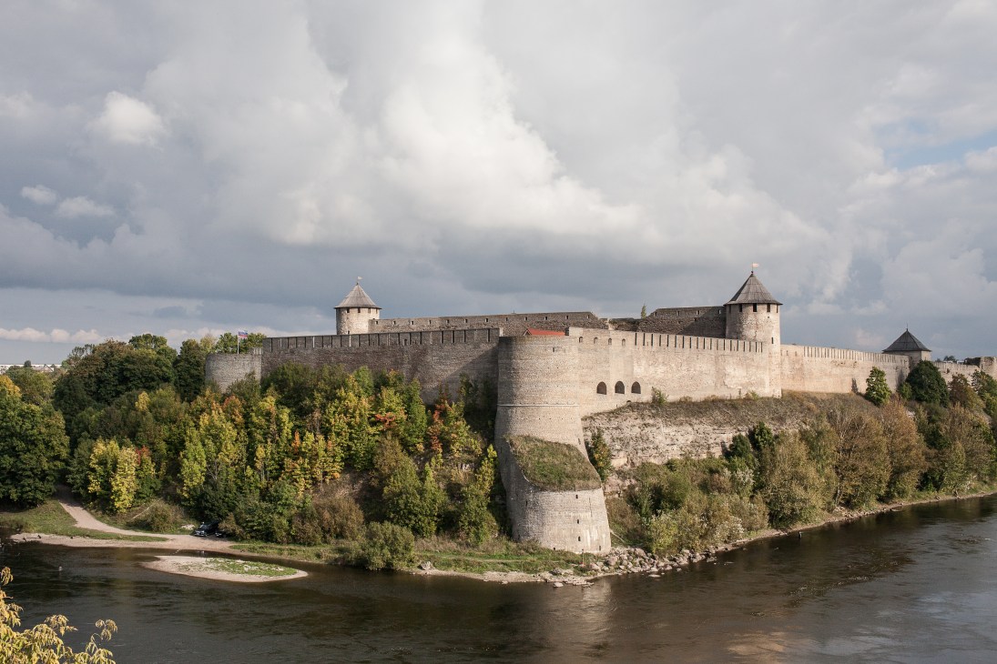 Medieval fortress with stone walls and towers on a forested riverbank under cloudy sky.