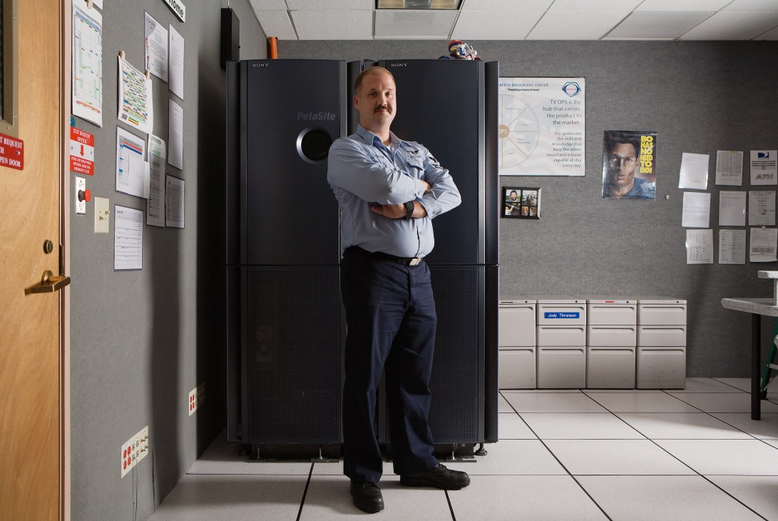 Petty officer Andrew Wiskow standing in front of large Sony broadcast monitors in a technical control room.