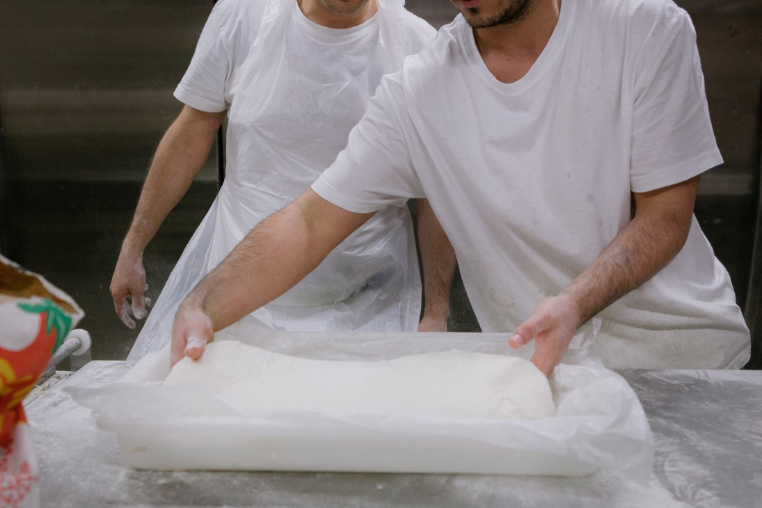 Two chefs in white shirts knead and stretch sourdough dough on a work surface.