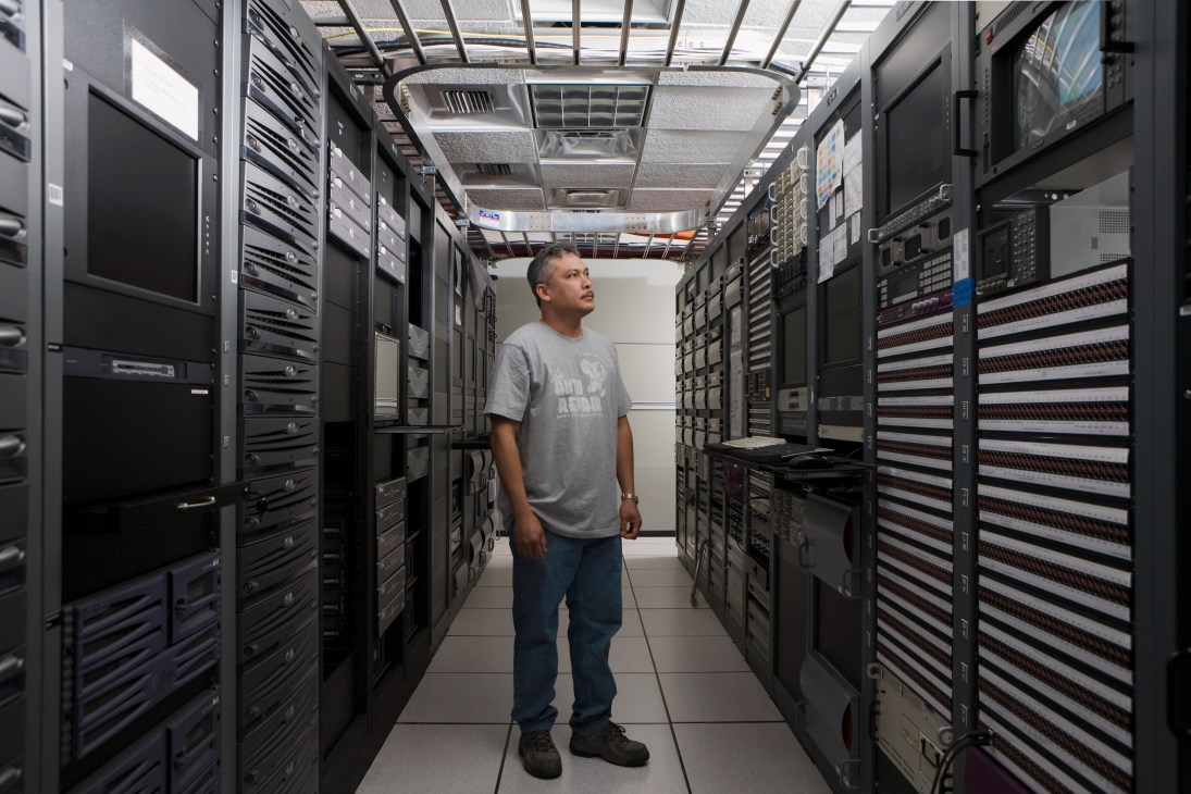 Broadcast engineer Jessie Balos standing in a server room lined with black equipment racks.