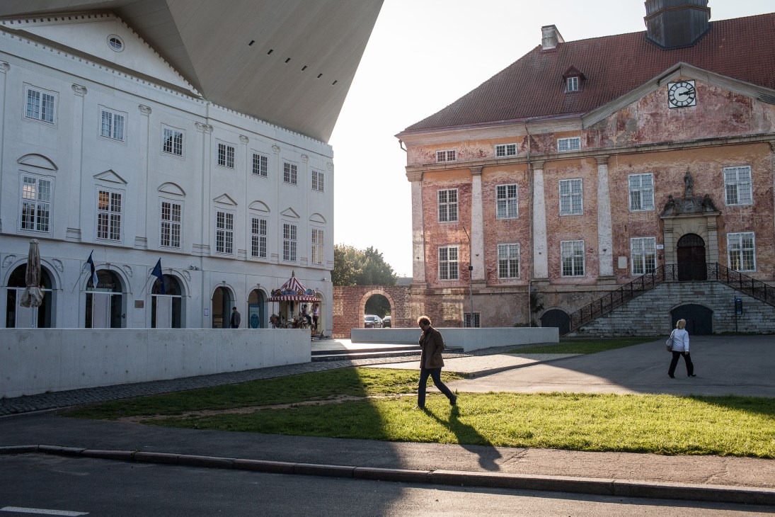 Two historic buildings in a town square: a white neoclassical college building on the left and a red-brick town hall with a clock tower on the right, with people walking across the grass between them.