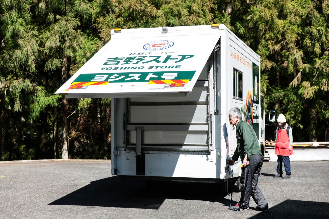 A white Yoshino Store mobile grocery truck with its expandable cabin extended, parked in a mountain area with customers nearby.