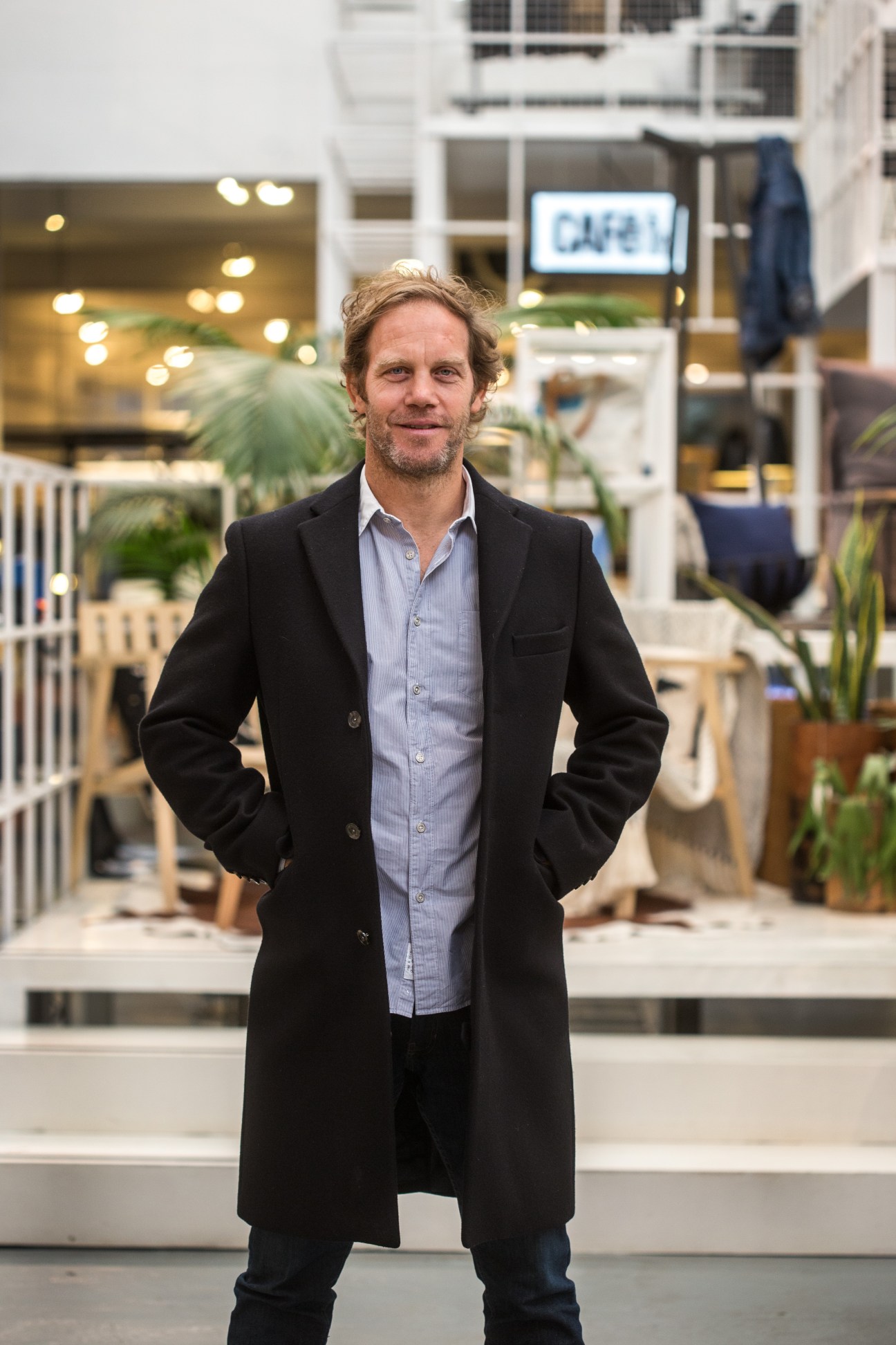 Man in black coat and light blue shirt standing in modern retail space with plants and signage.