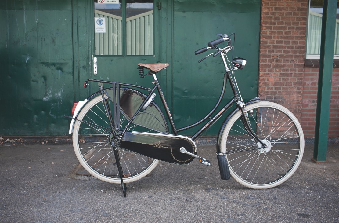 A dark blue Gazelle bicycle with brown leather saddle and integrated lock stands against a green door.