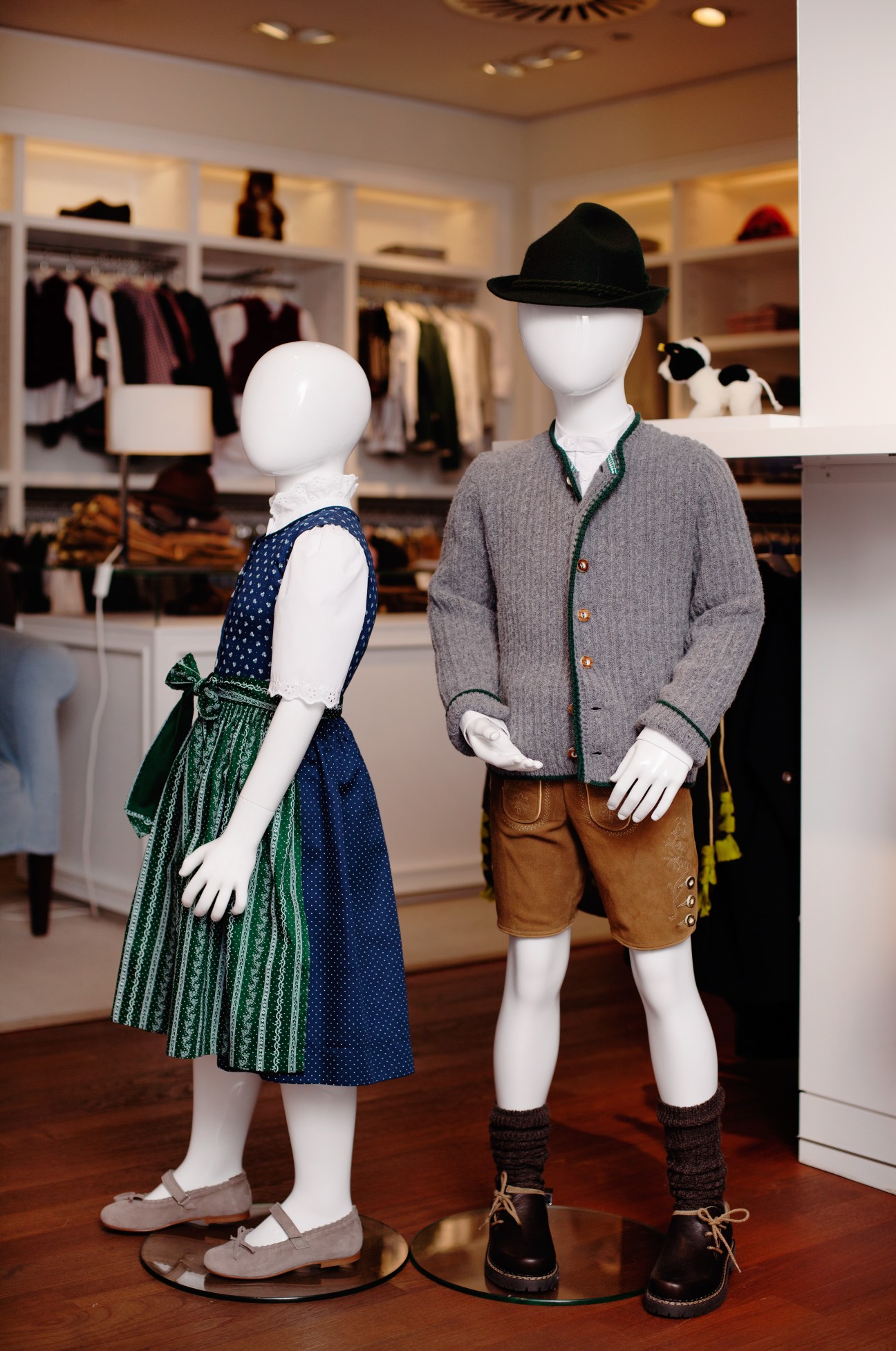 Two mannequins displaying children's traditional Bavarian tracht clothing at Lodenfrey, including a dirndl with green apron and lederhosen with hat.