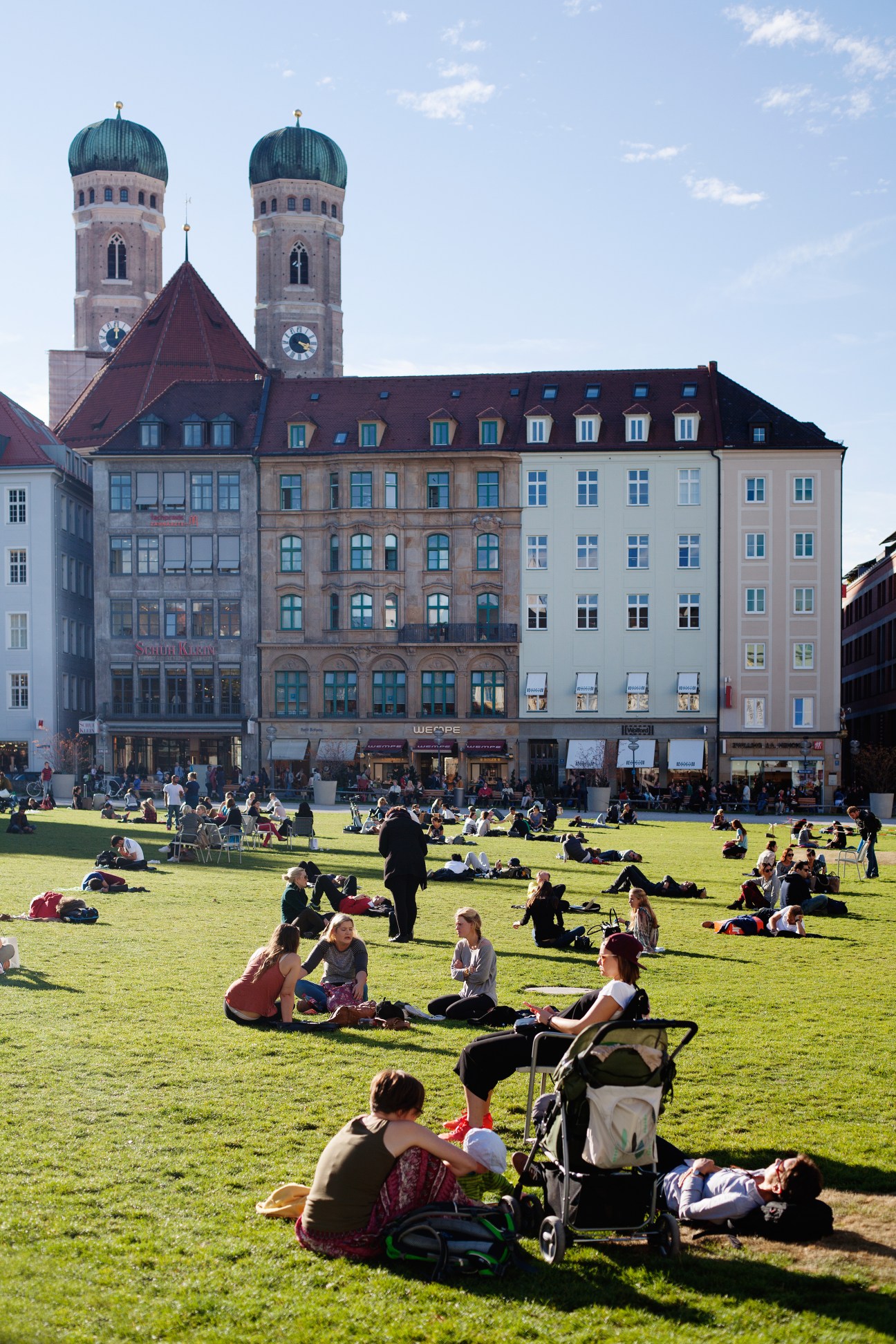 Downtown Munich square with historic buildings and twin-domed church towers in background, people relaxing on grass.