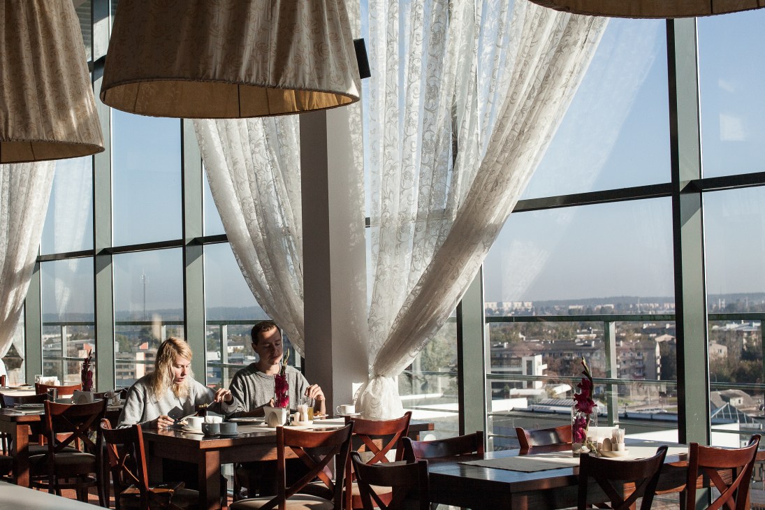 Two diners at a table in a high-rise restaurant with floor-to-ceiling windows overlooking Daugavpils cityscape.