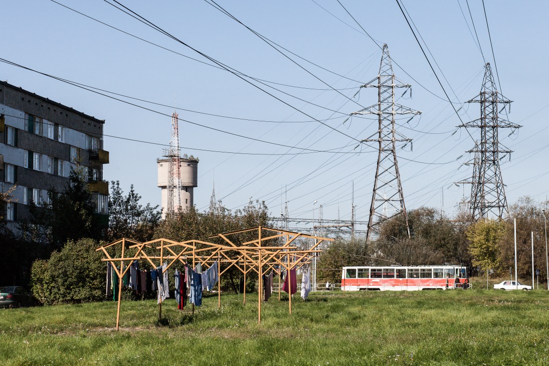 Clotheslines with laundry strung between wooden frames in a field, with Soviet-era apartment block, water tower, electrical pylons and red and white tram in background.