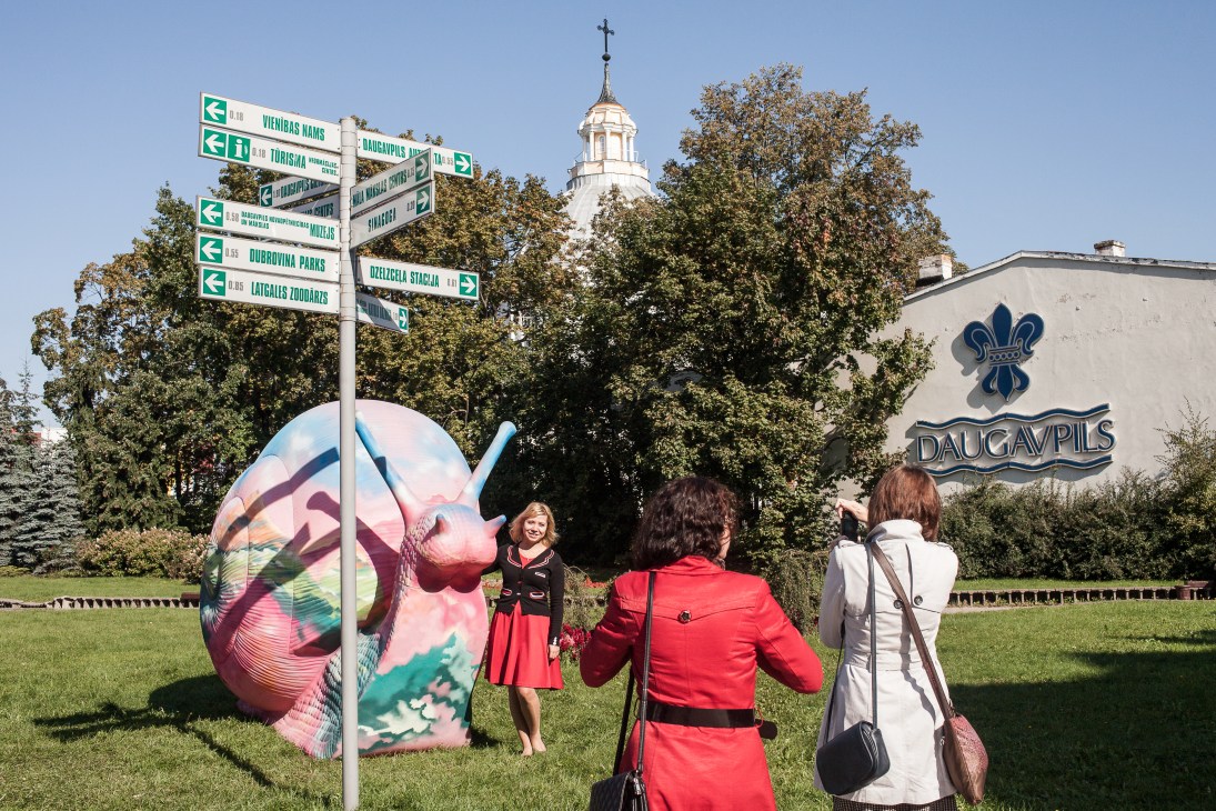 Visitors posing with a colorful abstract sculpture near directional signs in Daugavpils, Latvia.
