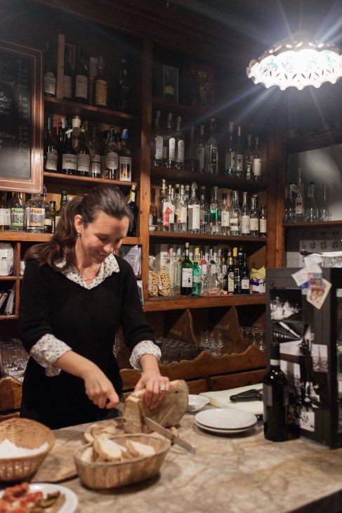 Woman serving food at wine bar Al Vino Al Vino in Rome's Monti neighbourhood, with wooden shelves of wine bottles behind the counter.