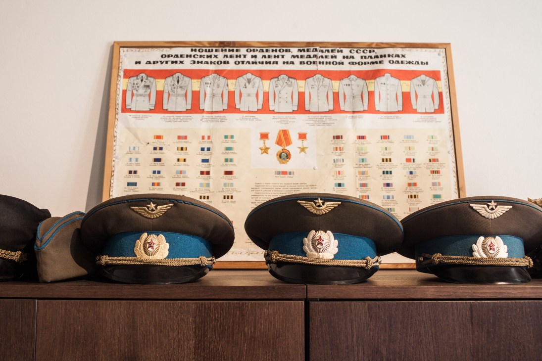 Soviet-era military peaked caps with gold insignia displayed on wooden shelf below framed uniform and medal chart.
