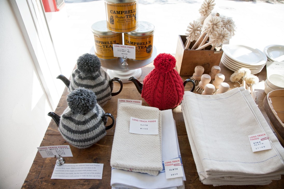 Long Track Foods shop display with Campbell's tea tins, knitted teapot cozies, cloths and dried flowers on wooden table.