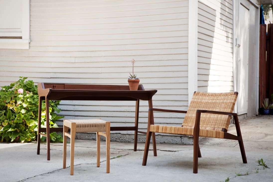 Mid-century modern furniture in an outdoor setting including a wooden desk, woven chair, and stool.