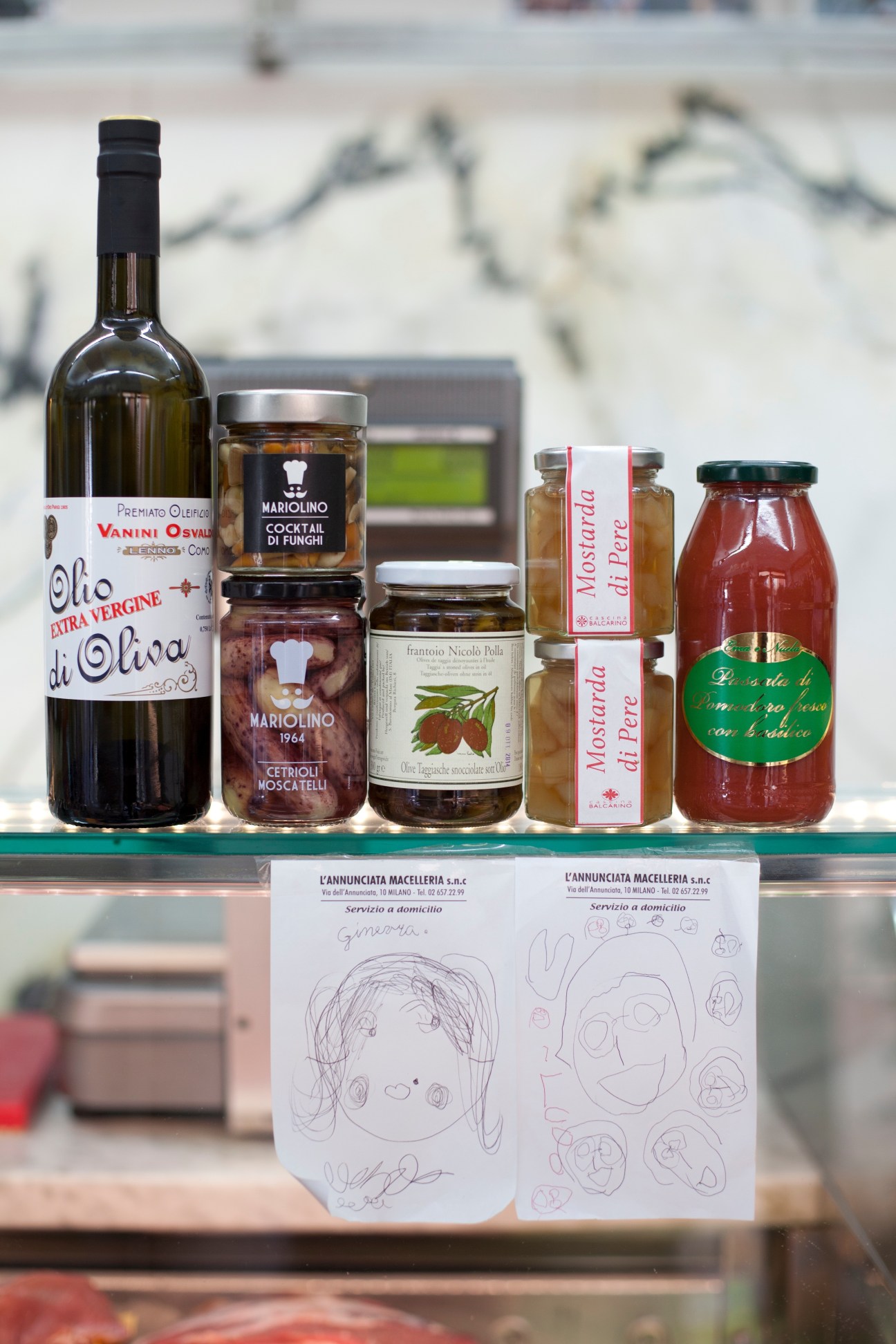 Italian specialty food products displayed in window including olive oil bottle, preserved mushrooms, and jars with handwritten children's drawings below.