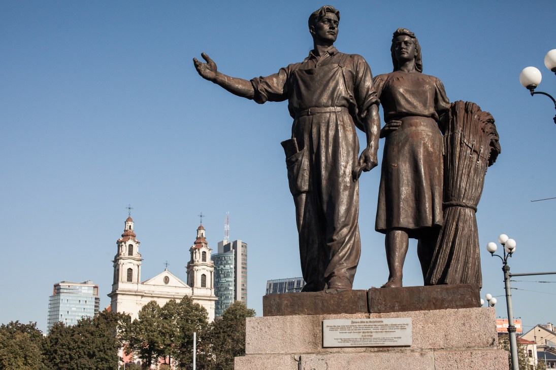 Bronze Soviet-era statues of two figures on a pedestal with a church and modern buildings behind them in Vilnius.