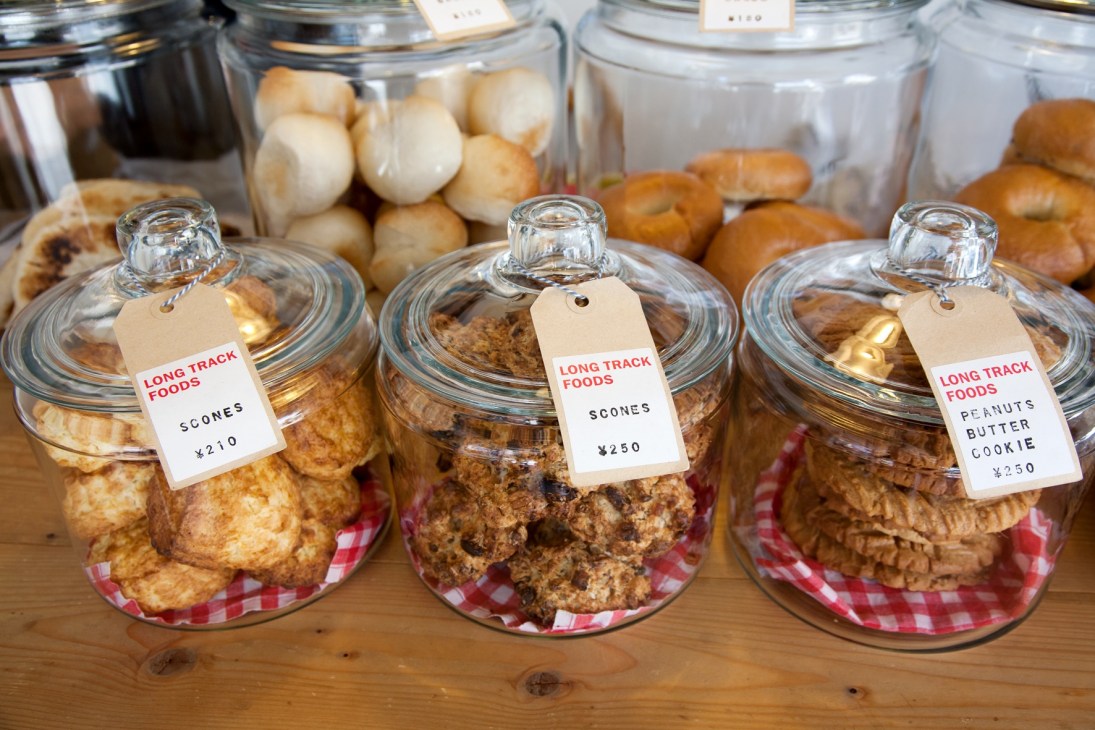 Glass jars filled with baked goods like scones and cookies labeled with Long Track Foods price tags on wooden counter.