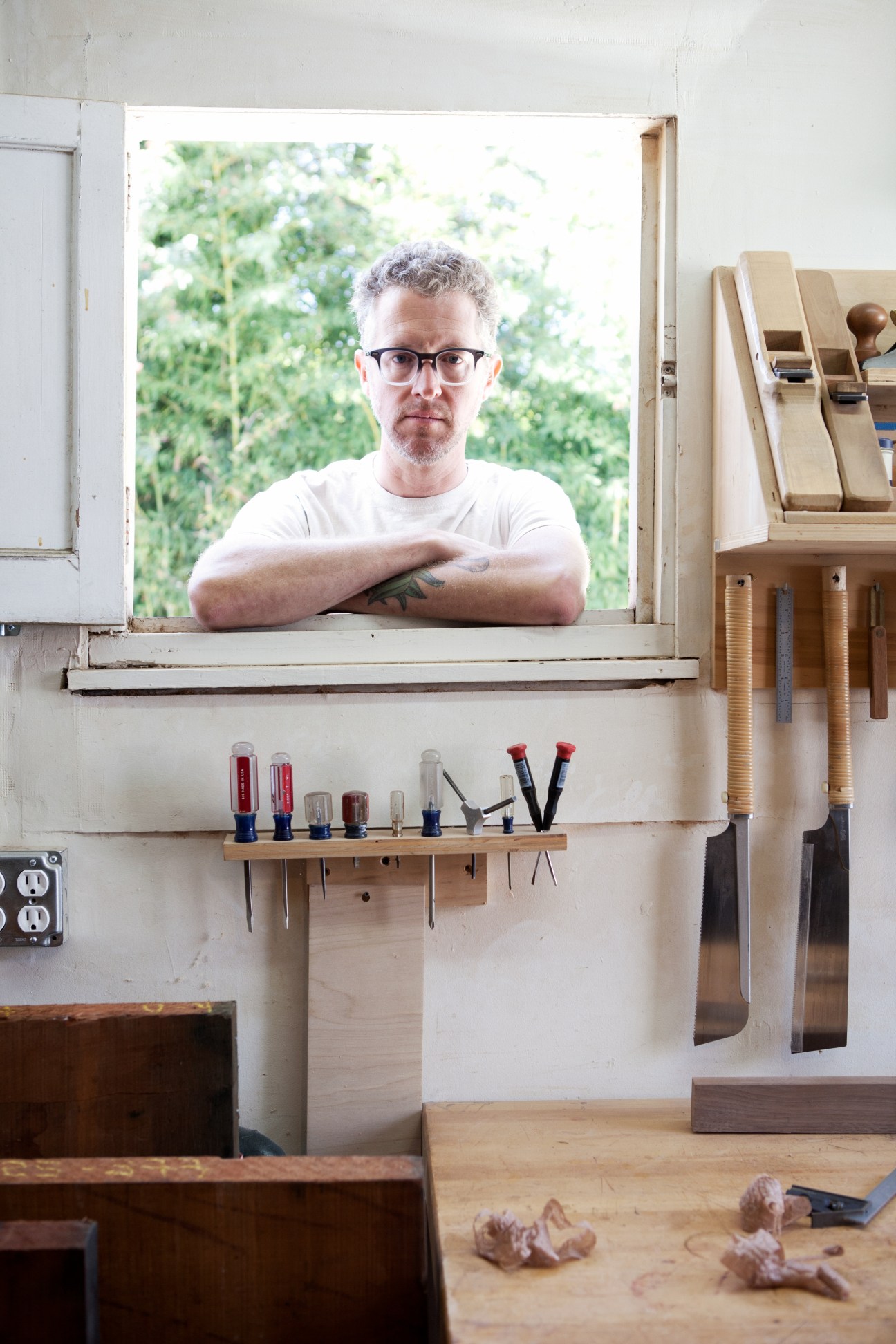Man with glasses standing at workshop window surrounded by woodworking tools and wooden furniture pieces.