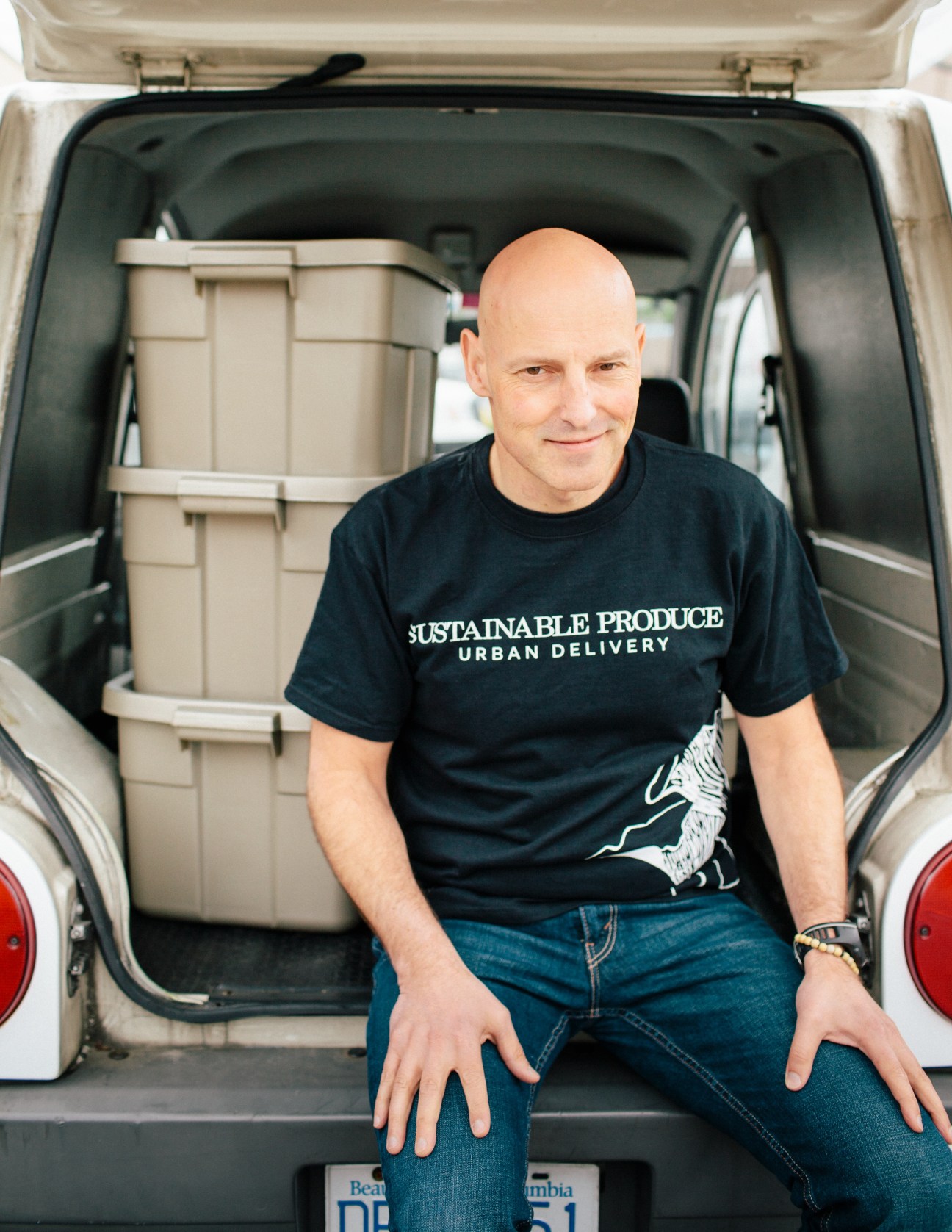 Man wearing SPUD shirt sits in open delivery van with produce boxes behind him.