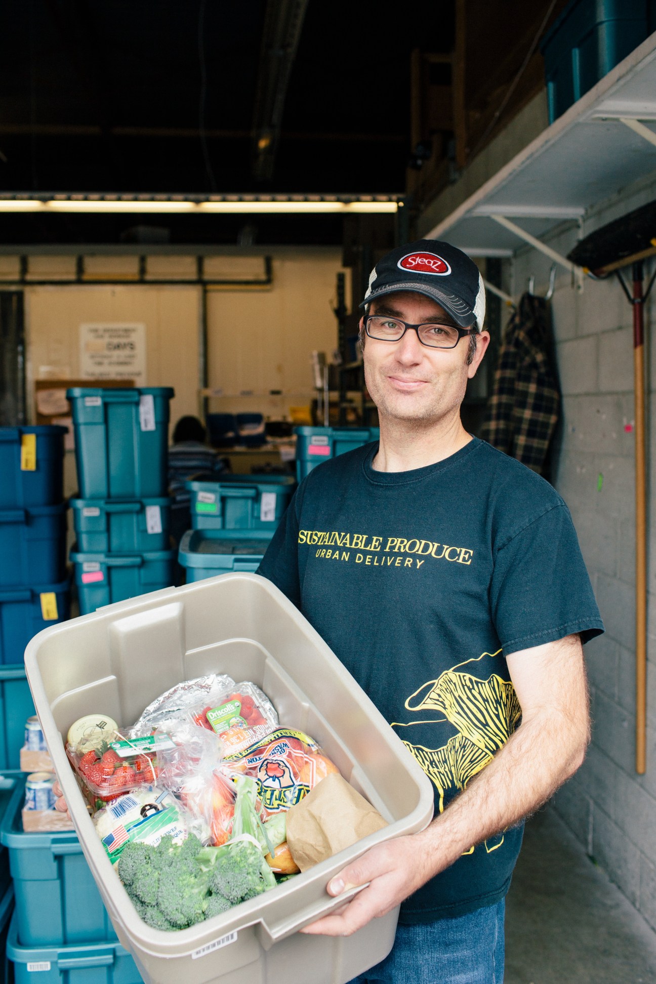 Man wearing SPUD t-shirt holding white delivery container with fresh produce in warehouse.