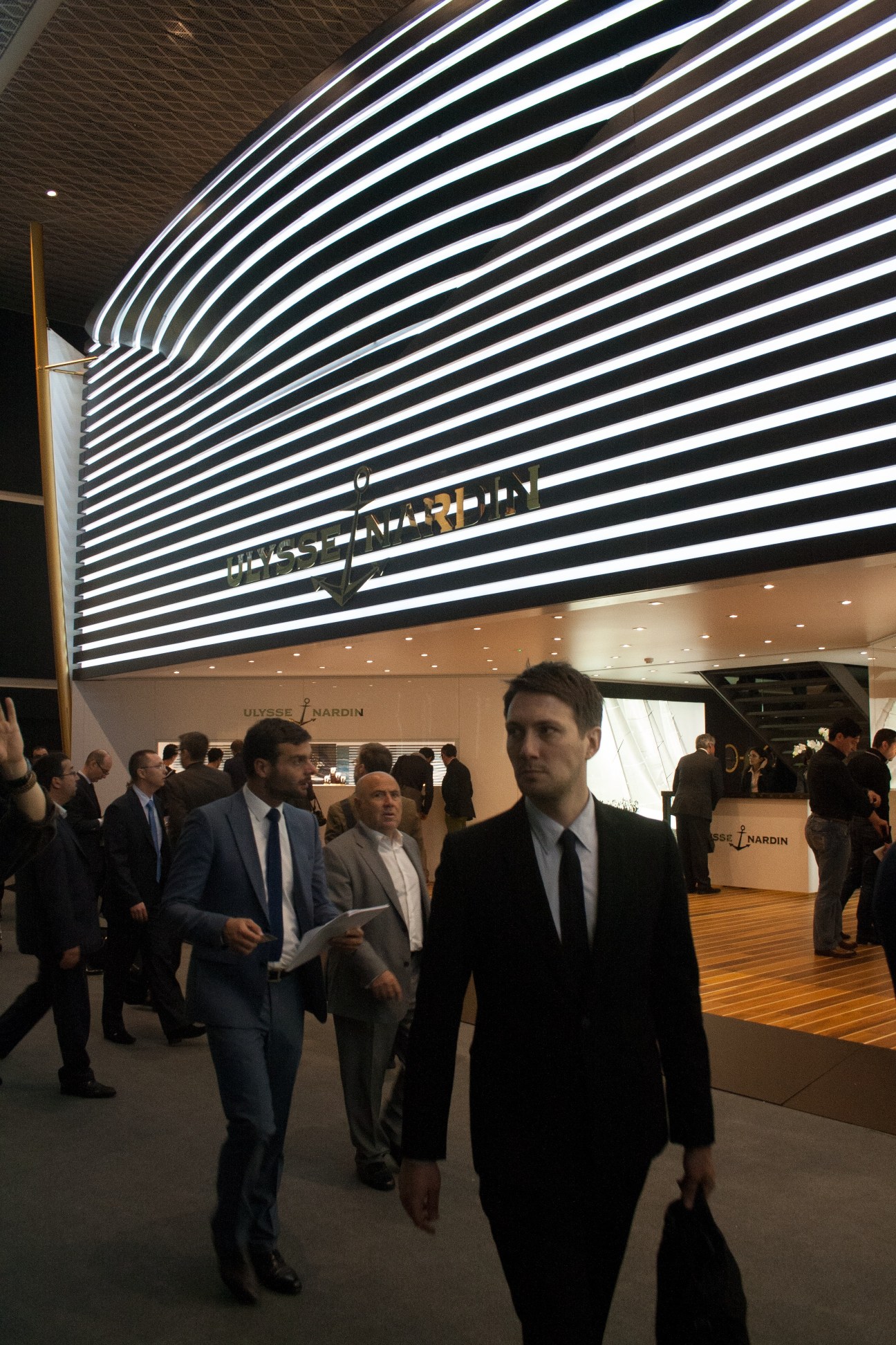 Visitors in business attire walking through Baselworld fair past the illuminated Ulysse Nardin booth with striped architectural feature.